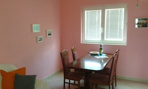 Een eethoek met een tafel, stoelen, een raam met jaloezieën en enkele ingelijste foto's aan de muur.
