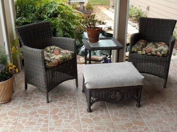 A patio area with wicker furniture, including two chairs and a small table, surrounded by potted plants and a stone-tiled floor.