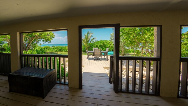 A wooden deck with a table and chairs, a trash bin, and a view of a pool and greenery. - Corktree Beach House - Modern, Beachfront 2bd/3ba home with central AC (Grand Turk)