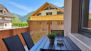 Una terraza de madera con una mesa, sillas, una planta en maceta y vistas a una montaña y casas al fondo.