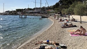 A beach with people sunbathing and swimming, a pier with boats, and buildings in the background. - VILLA NARESTE Stone house with pool and amazing sea view (Jesenice)