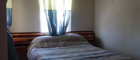 Une petite pièce à la lumière tamisée, avec un lit simple, une tête de lit en bois et une fenêtre ornée de rideaux légers.