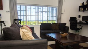 Sala de estar con sofá, sillón y mesa de centro. Se ve un paisaje verde a través de la ventana.