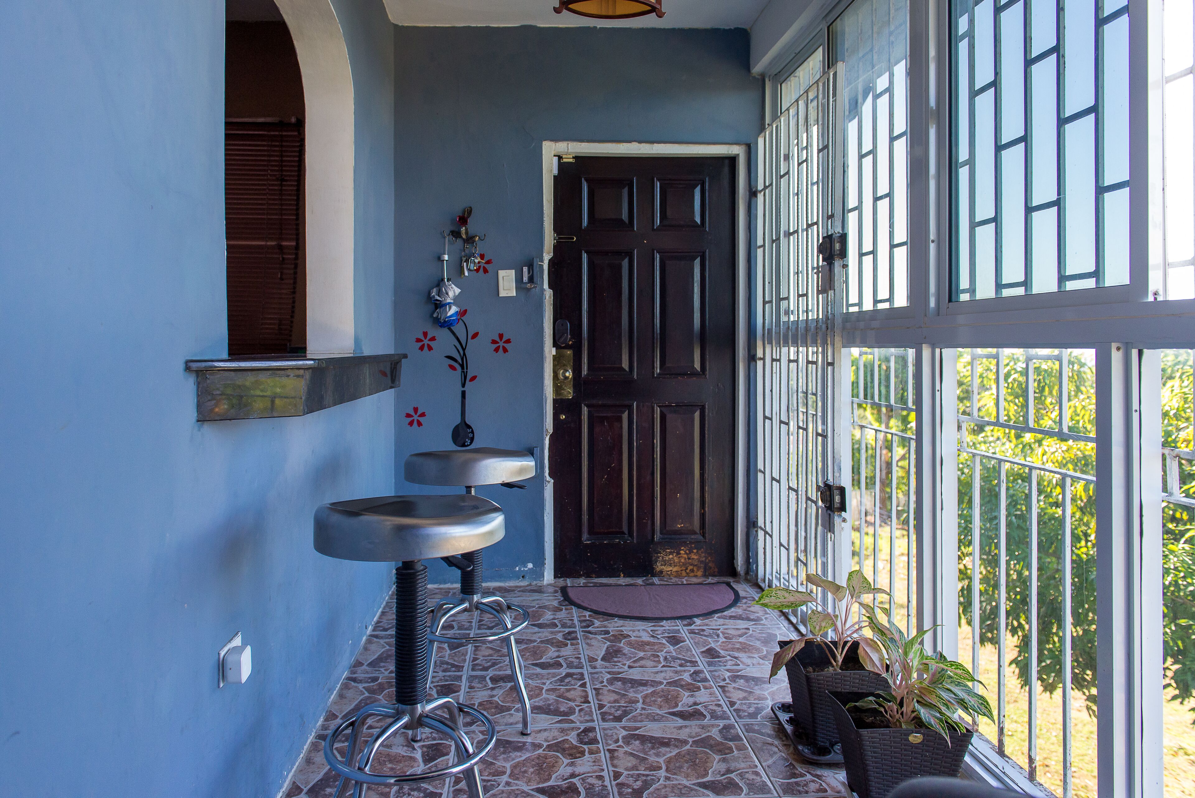 Un couloir avec un mur bleu, une porte en bois foncé et une fenêtre ornée d’un garde-corps en métal. On y trouve deux tabourets de bar et une plante en pot près de la porte.