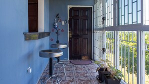 Un pasillo con pared azul, una puerta de madera oscura y una ventana con barandilla metálica. Hay dos taburetes y una planta en maceta cerca de la puerta.