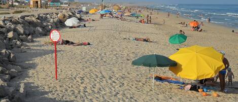 Praia com pessoas se banhando e tomando sol, placa de proibição de banho e guarda-sóis.