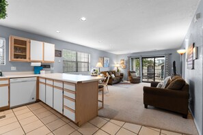 A modern kitchen with a central island, a dining area with a table and chairs, a sofa, and a large sliding glass door leading to an outdoor patio.