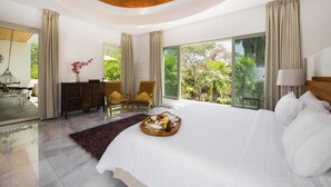 Un dormitorio espacioso con una cama grande, un área de comedor con una bandeja de desayuno y vistas a abundante vegetación a través de amplias ventanas.