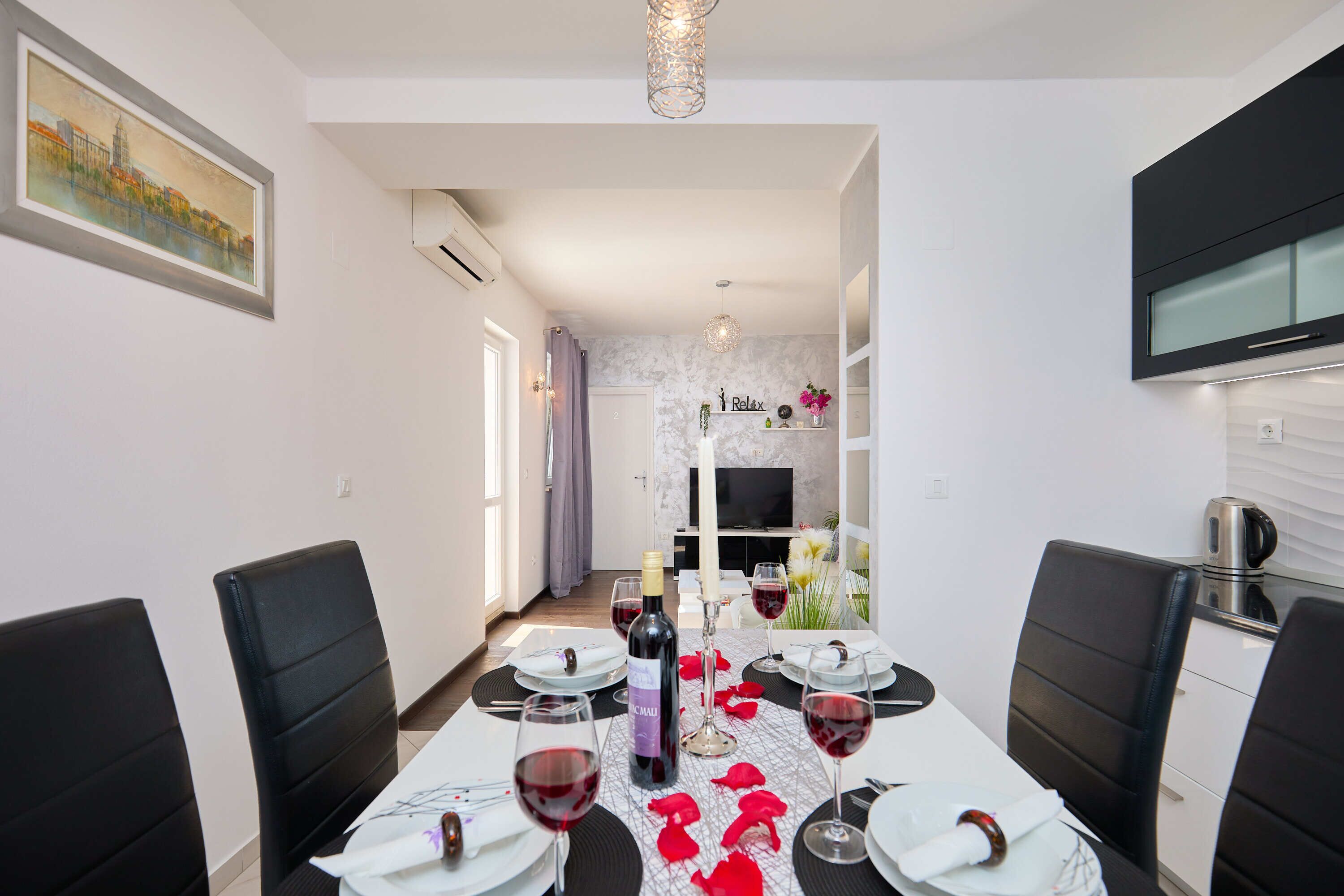 A dining area with a table set for four, a bottle of wine, and red rose petals on the table.