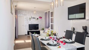 Uma sala de jantar moderna com uma mesa comprida posta para quatro pessoas, uma televisão de tela plana na parede e uma área de cozinha com uma máquina de café.