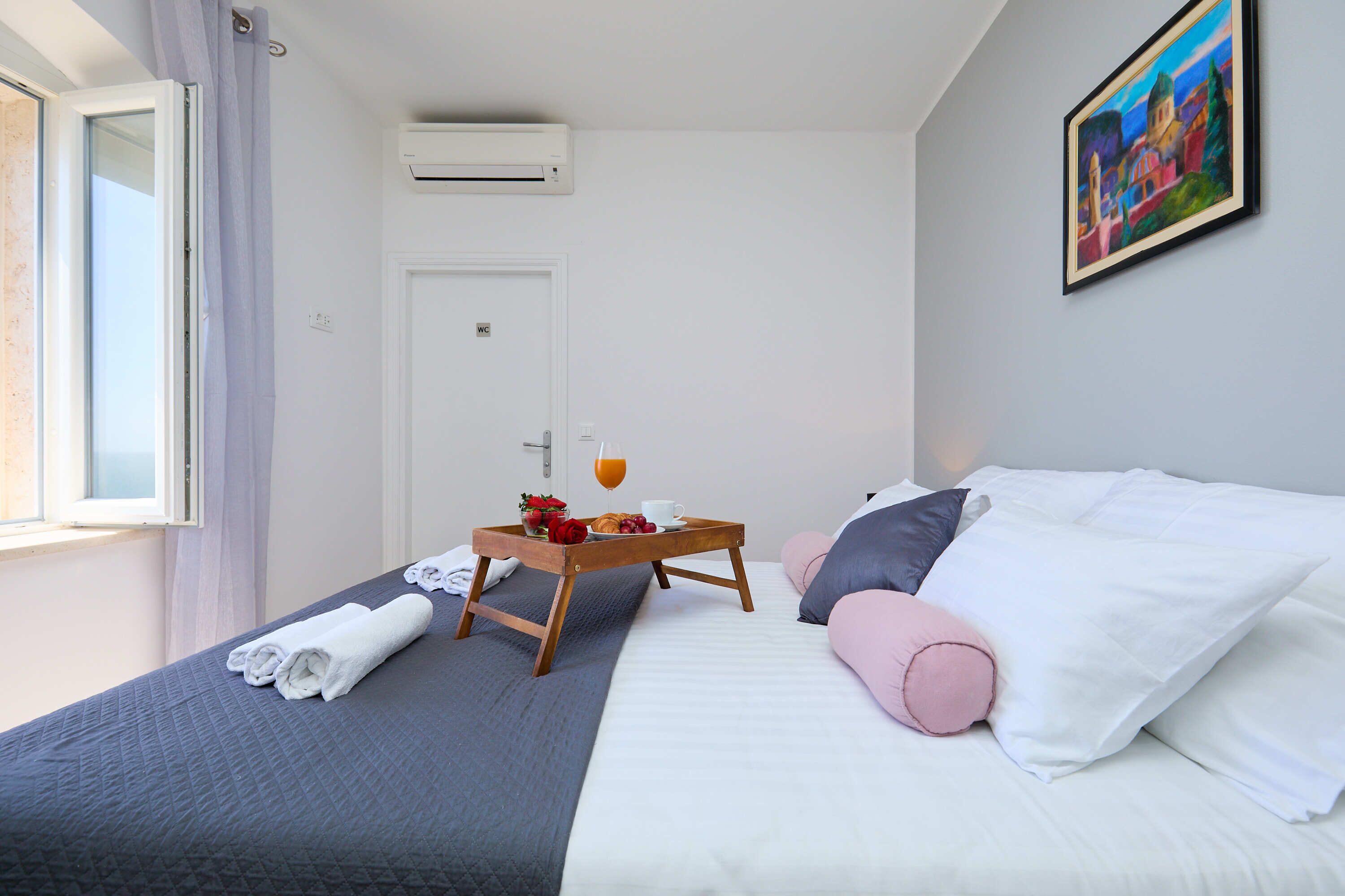 A hotel room with a bed, a bedside table with a tray of breakfast, a window with curtains, and an air conditioning unit.
