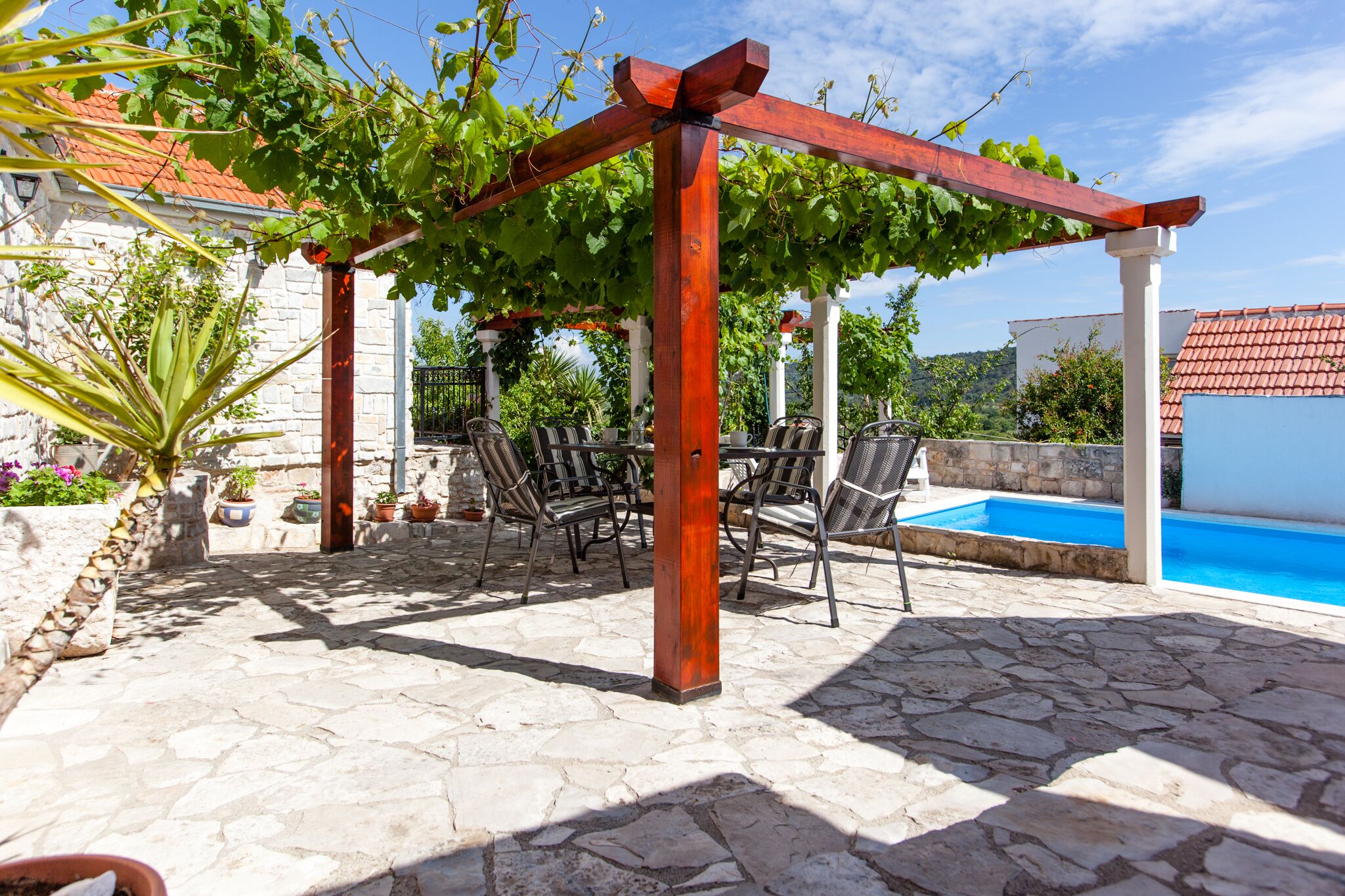 A stone patio with a pergola covered in green vines, outdoor seating, and a swimming pool.