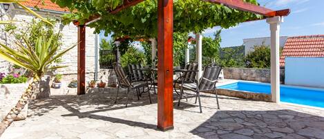 A stone patio with a pergola covered in green vines, outdoor seating, and a swimming pool.