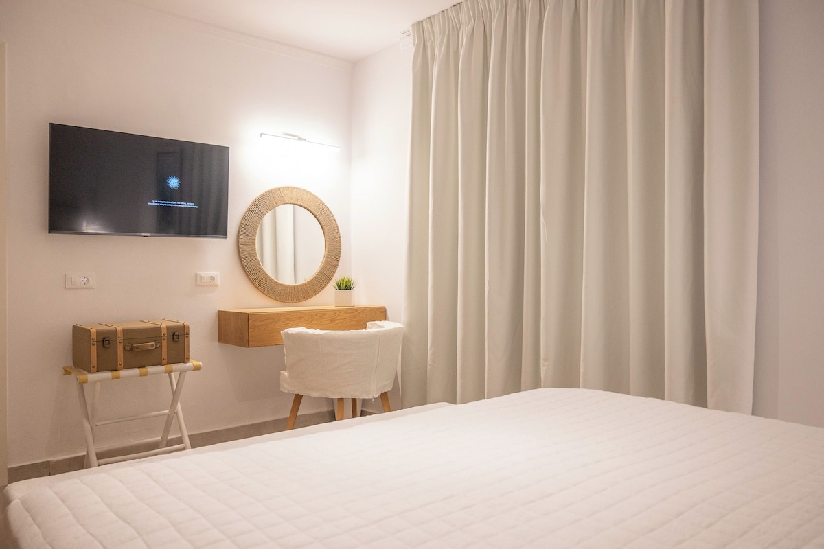 Habitación de hotel con cama, televisor montado en la pared, escritorio de madera con espejo, maleta y una silla blanca.