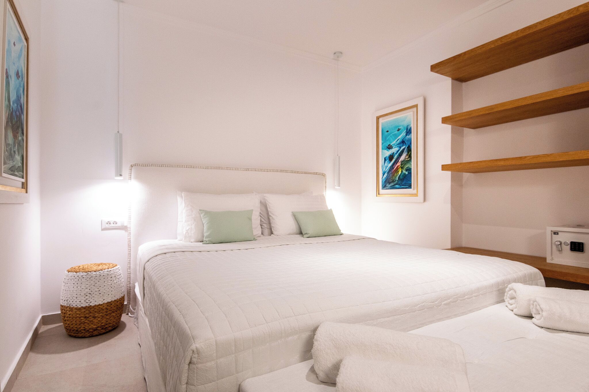 A bedroom with two beds, a bedside table, a wall-mounted shelf, and a painting.