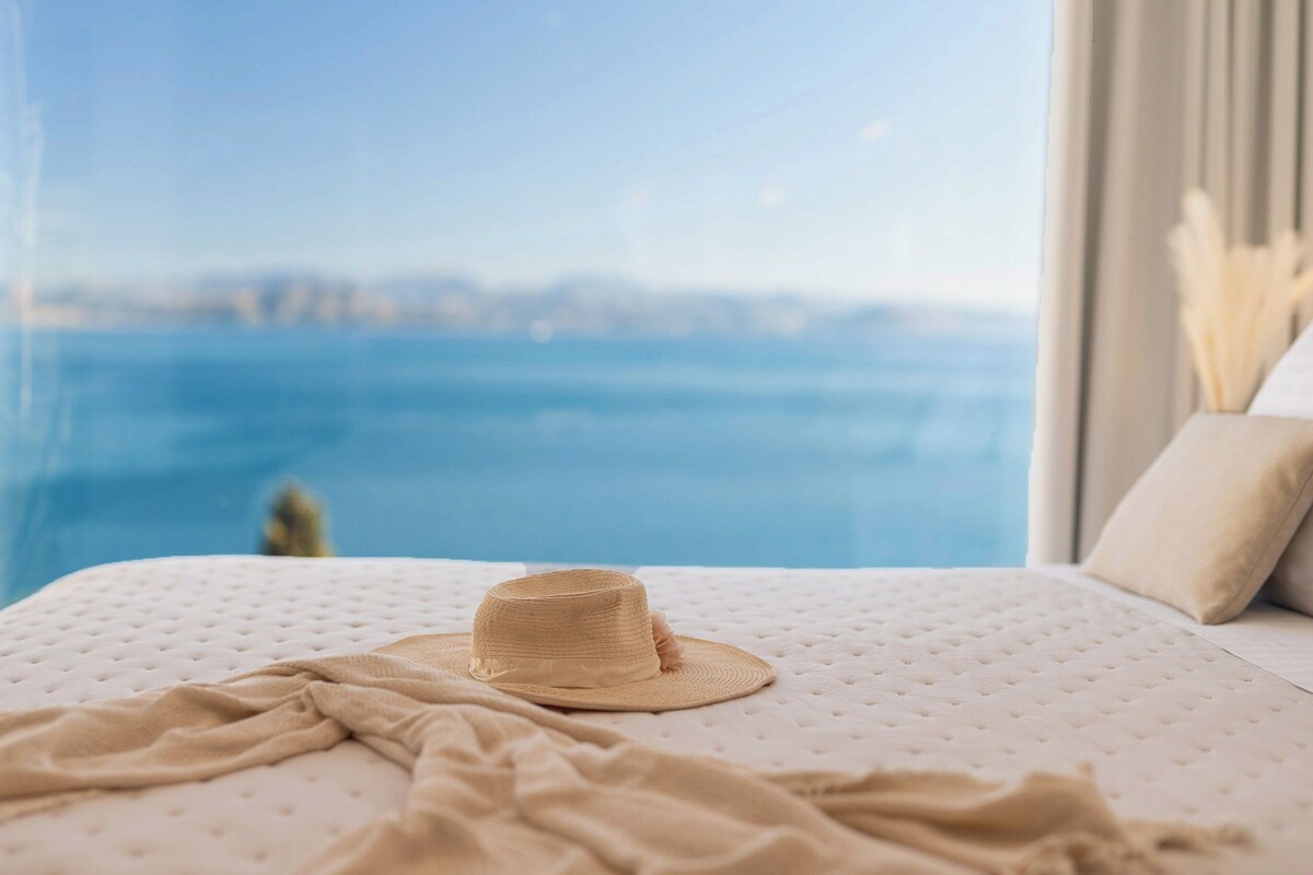 A bed with a beige blanket and a straw hat, a window with a view of the sea, and a beige pillow.