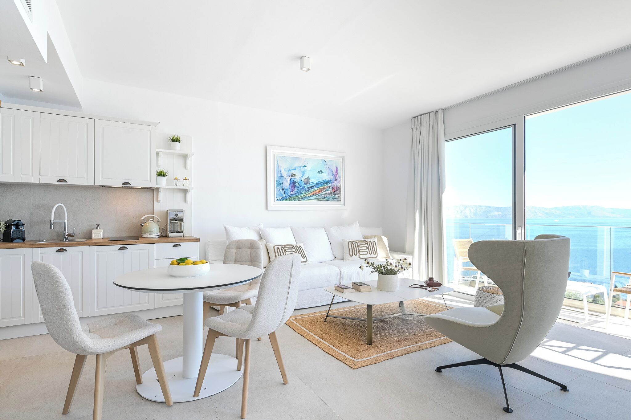 A modern, minimalist living room with a dining area, white furniture, and a large window with a view.