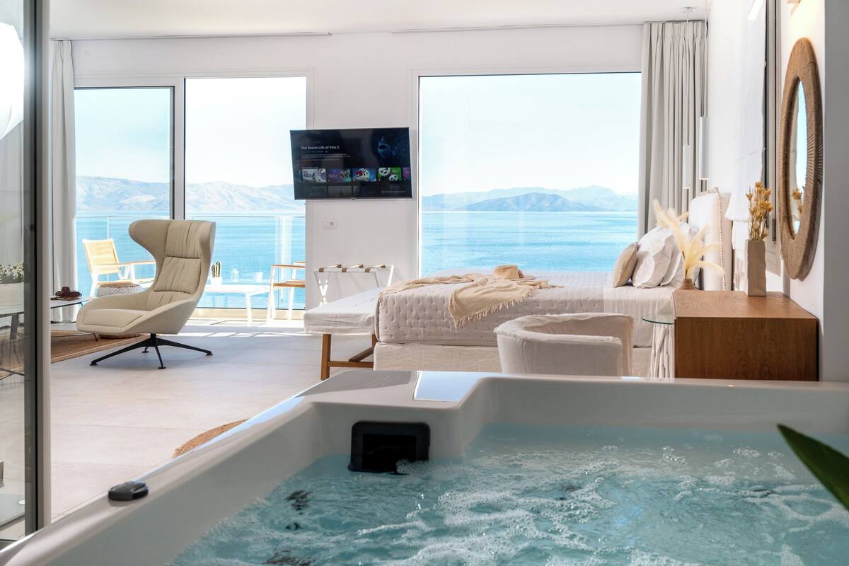 A modern hotel room with a large window, a bed, a sofa, a TV, and a hot tub.