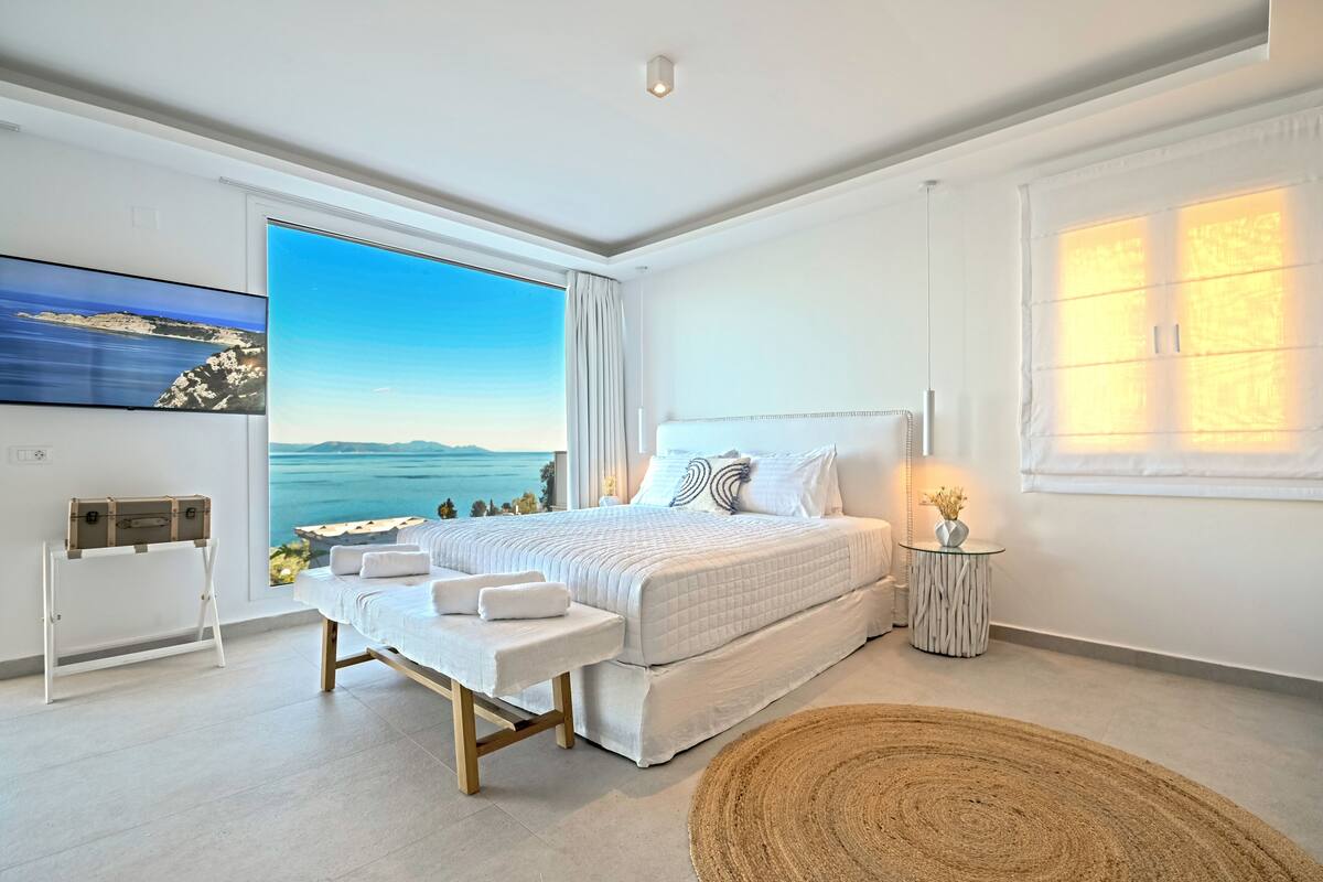 A modern bedroom with a large bed, a view of the sea, a small bedside table, and a large window.