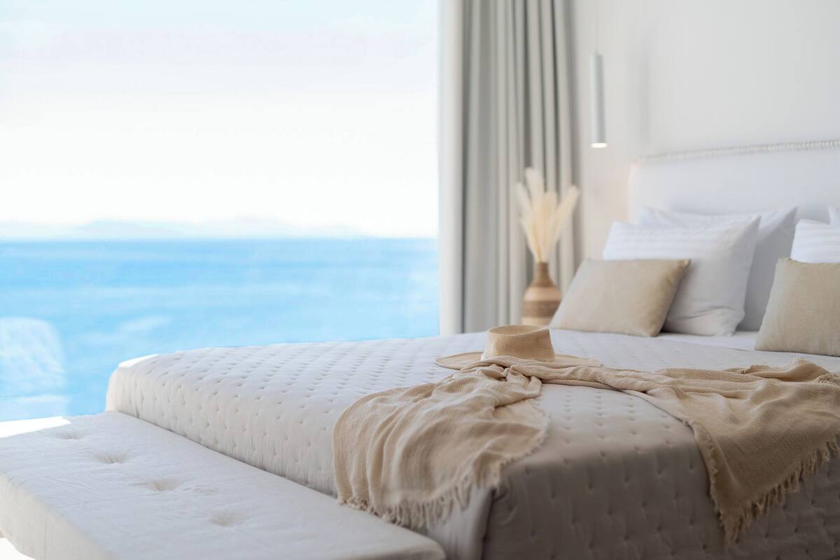A bedroom with a large window, a bed with a beige blanket, and a vase with dried flowers.