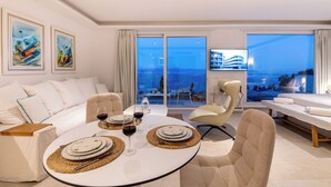 Un salon moderne avec une table à manger ronde pour quatre personnes, un canapé et une table de massage. De grandes fenêtres offrent une vue sur la mer et un immeuble.
