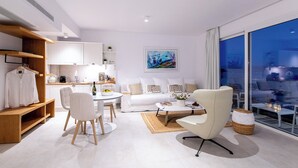 Un salon moderne avec un canapé blanc, une table basse et un coin repas comprenant une table et des chaises blanches. Une grande fenêtre offre une vue sur la ville.