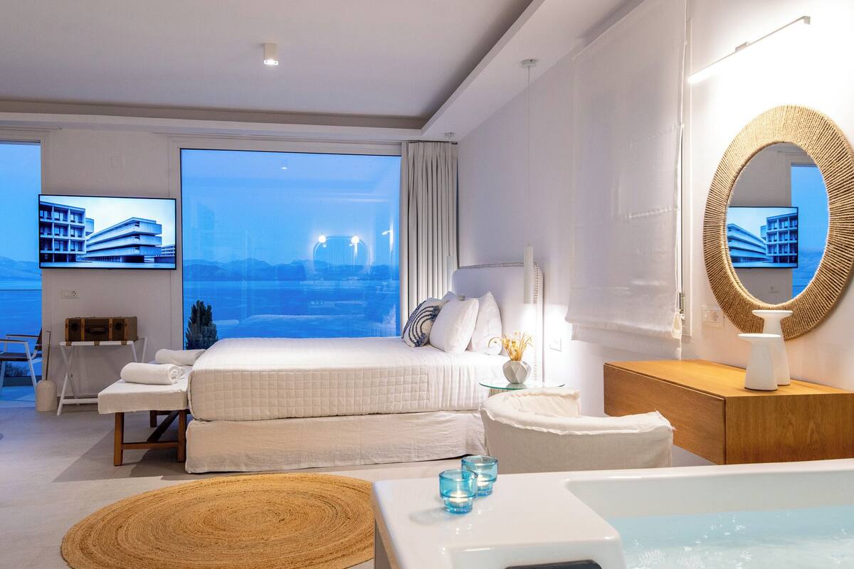 A modern hotel room with a large bed, a sitting area, a bathroom with a bathtub, and a view of the cityscape.