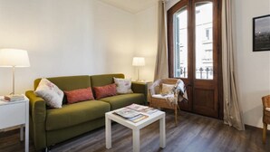 Una sala de estar con un sofá verde, dos mesitas de noche con lámparas, un sillón de mimbre, una puerta de madera y un cuadro en la pared.