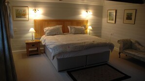 A bedroom with a wooden headboard, two bedside lamps, a floral bedspread, a small bedside table, a chair, and framed pictures on the walls. - Converted Granary in SE Kent UK (Lyminge, nr Folkestone)