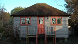 A small, single-story wooden house with a red door and windows, surrounded by vegetation. - Converted Granary in SE Kent UK (Lyminge, nr Folkestone)
