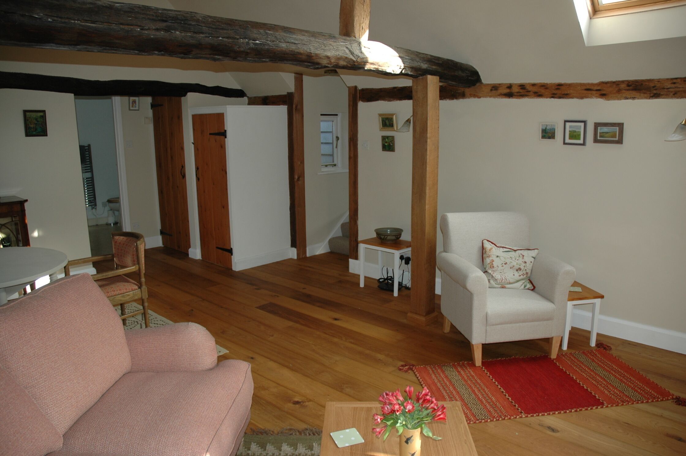 Un salon avec un plancher en bois, un canapé, un fauteuil, une petite table sur laquelle est posé un vase de fleurs et un tapis rouge.