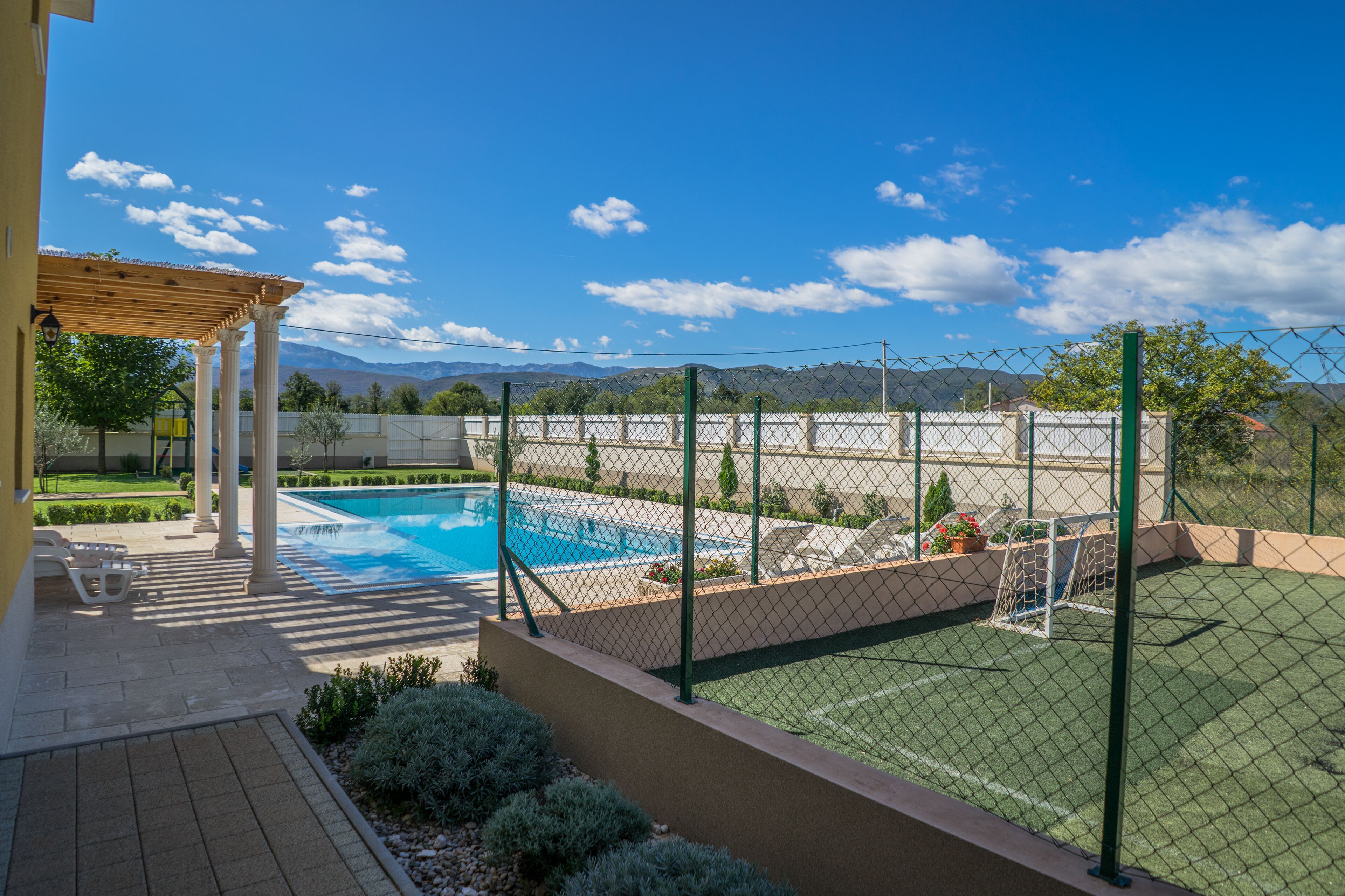 A swimming pool with a pergola, a fenced tennis court, and a view of mountains.