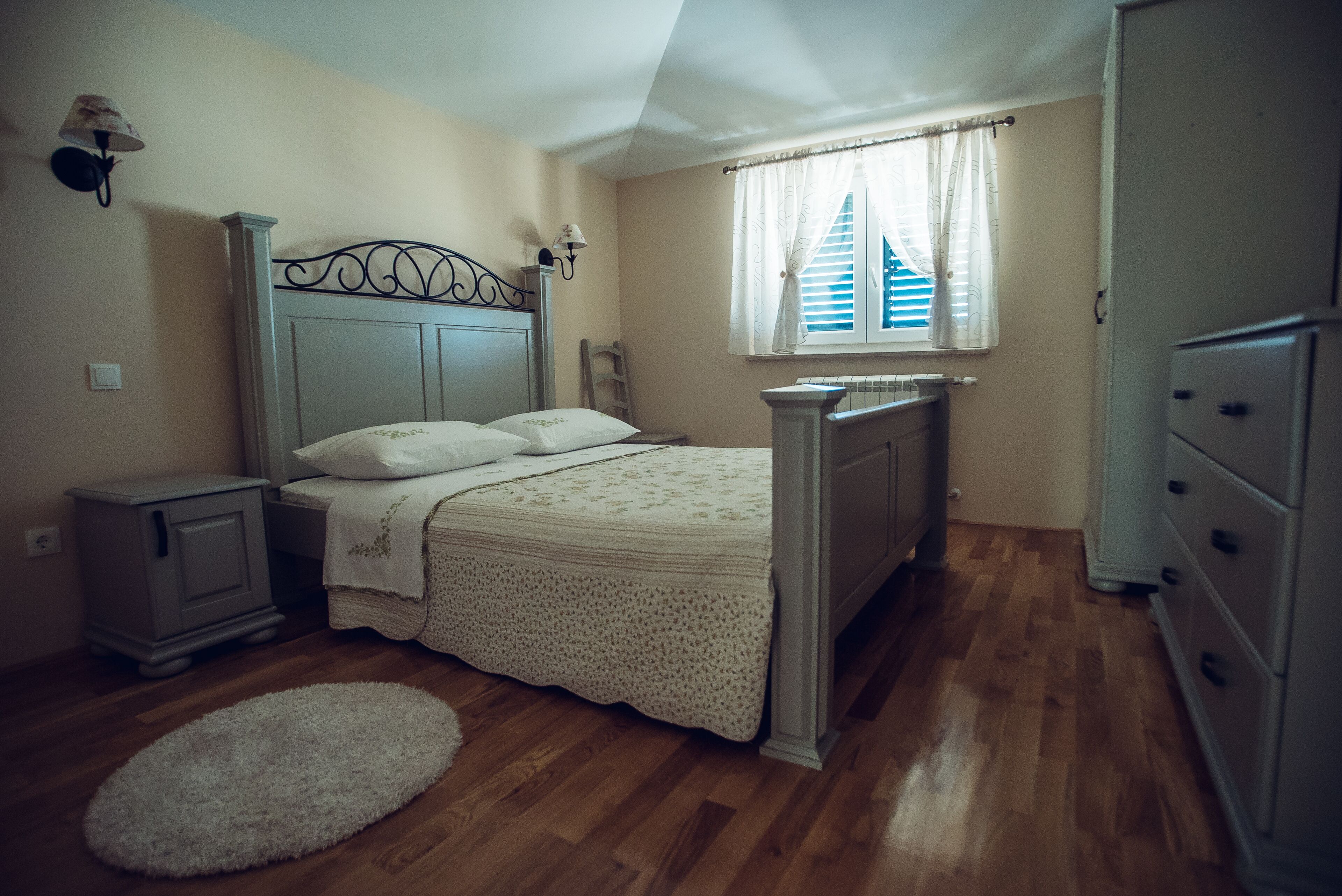 A bedroom with a bed, a nightstand, a dresser, a window with curtains, and a radiator.