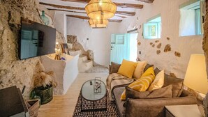 A rustic room with exposed beams, a sofa with cushions, a hanging light fixture, and a television mounted on the wall. - Cave Suite THA (Cave Hotel Lomas de Ruvira) (Jorquera)
