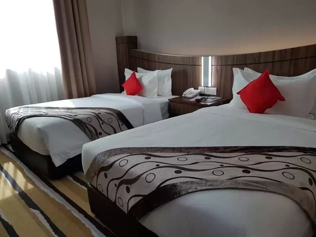 Quarto de hotel com duas camas, cada uma com colcha estampada, cabeceira de madeira e mesa de cabeceira com telefone e almofada vermelha.
