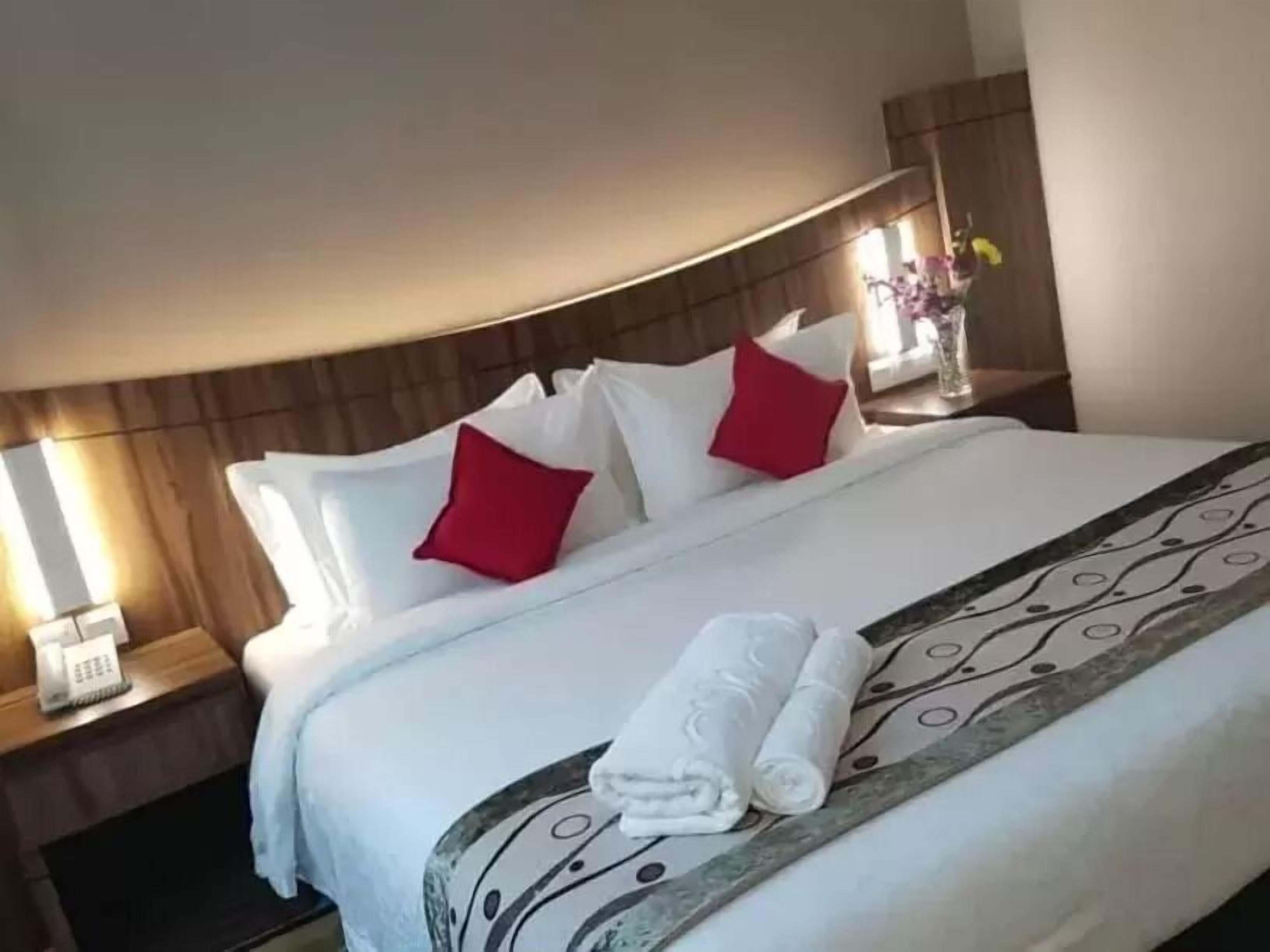 A neatly made bed with white linens, red pillows, and a rolled towel.