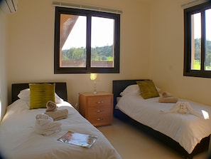 Two single beds with dark headboards and white bedding, a nightstand with a lamp, and large windows offering a view of the surrounding greenery.