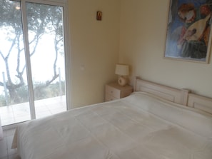 A bedroom with a large bed, a bedside table with a lamp, a painting on the wall, and a window with a view of trees.