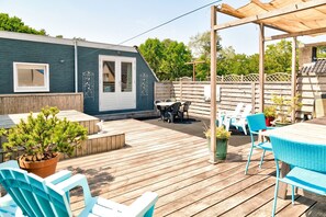 Una terraza de madera con mobiliario exterior, un pérgola y un pequeño patio con una mesa y sillas.
