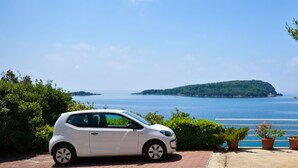 Ein weißes Auto steht auf einem befestigten Untergrund mit Blick auf ein Gewässer und eine entfernte Insel.