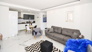 Ein modernes Wohnzimmer mit einem grauen Sofa, einem weißen Esstisch mit Stühlen und einer Küche im Hintergrund.