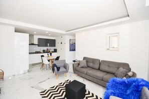 Una sala de estar moderna con un sofá gris, una mesa de comedor blanca con sillas, y una cocina al fondo.