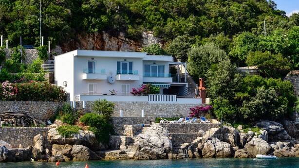 Ein modernes, zweistöckiges Haus mit Balkon, auf einem felsigen Hügel mit Blick auf ein Gewässer.