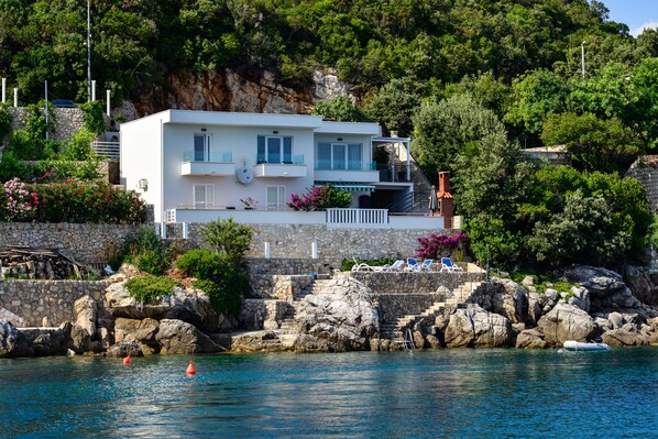 A modern two-story house with a balcony, situated on a rocky hillside overlooking a body of water.