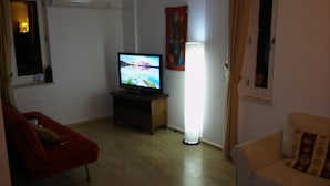A living room with a television on a wooden stand, a red armchair, a beige sofa, a standing lamp, and a window with curtains.