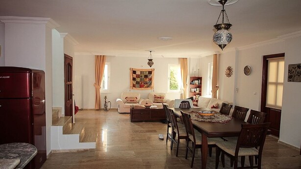 A spacious living area with a dining table, sofa, and a kitchen area with a refrigerator.