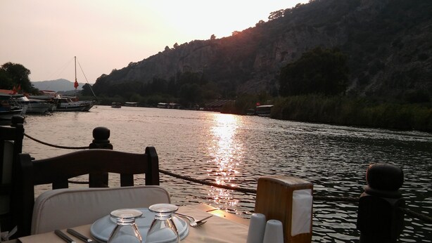 Tavola imbandita per la cena con vista su un fiume, barche e un paesaggio montuoso.