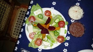 A plate of salad with tomatoes, cucumbers, onions, and cheese, accompanied by a bowl of rice and a small dish of beans. - Real family hostel, Hospedaje La Penita, 2 person room  (Altagracia)