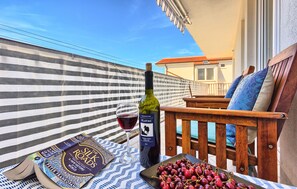 A balcony with a striped fabric backdrop, a wooden chair with cushions, a bottle of wine, a glass of red wine, a book titled 'The Silk Roads', and a plate of cherries.
