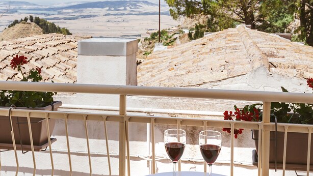 Una terraza con una mesa dispuesta para dos personas, con un cuenco de frutos secos y dos copas de vino tinto, con vistas a un paisaje pintoresco con montañas y árboles.